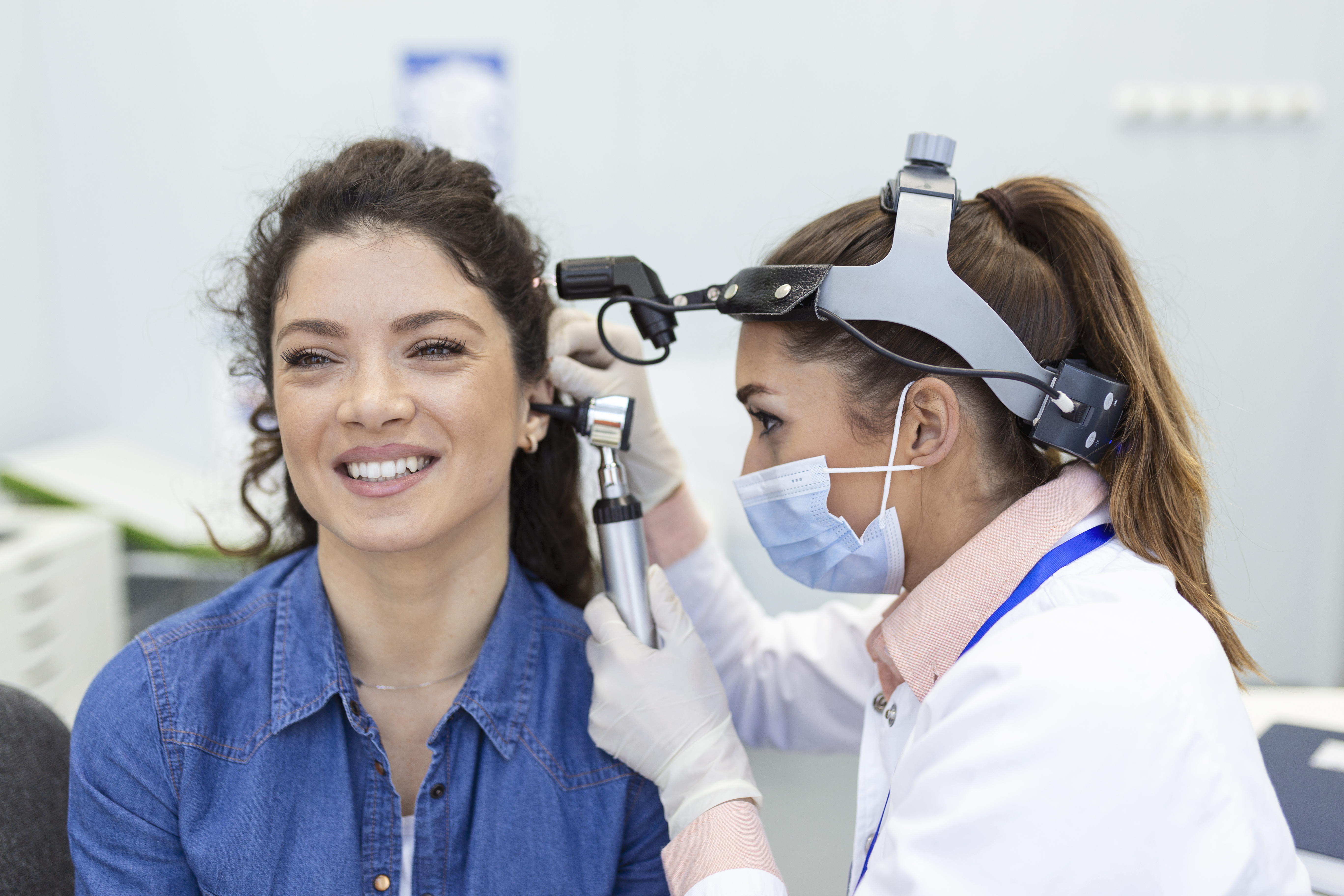 Picture of woman with brown hair in blue shirt smiling as the a doctor wearing a white lab coat and mask looks in the patients ear with an otoscope.