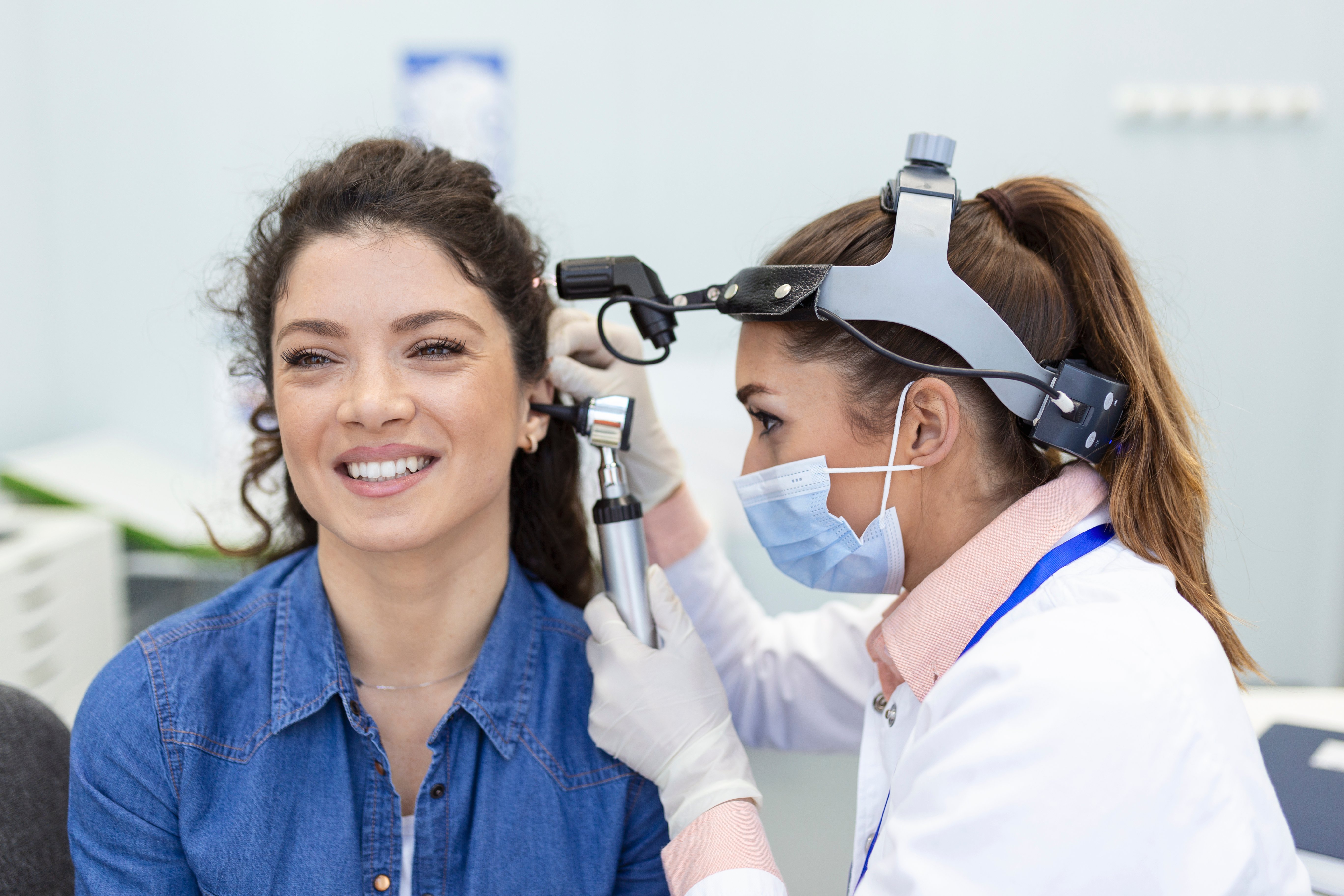 Picture of woman with brown hair in blue shirt smiling as the a doctor wearing a white lab coat and mask looks in the patients ear with an otoscope.